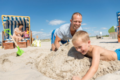 Lachende vader en zoon bouwen samen een groot zandkasteel op het strand, met een klassieke Duitse strandstoel op de achtergrond.