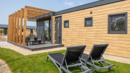 Buitenaanzicht van een modern houten chalet met een overdekt terras en twee ligbedden in de tuin op Strandpark De Zeeuwse Kust.