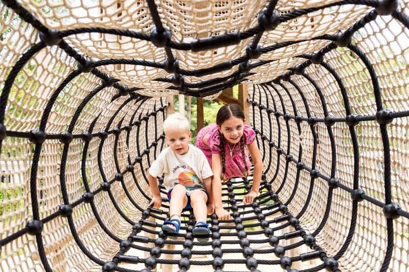 Twee kinderen klimmen door een tunnel van netten in de buitenspeeltuin van Bospark Ede op de Veluwe.