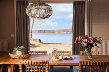 Eetkamer met een houten tafel gedekt voor een maaltijd en een groot raam dat uitkijkt op het strand en het meer