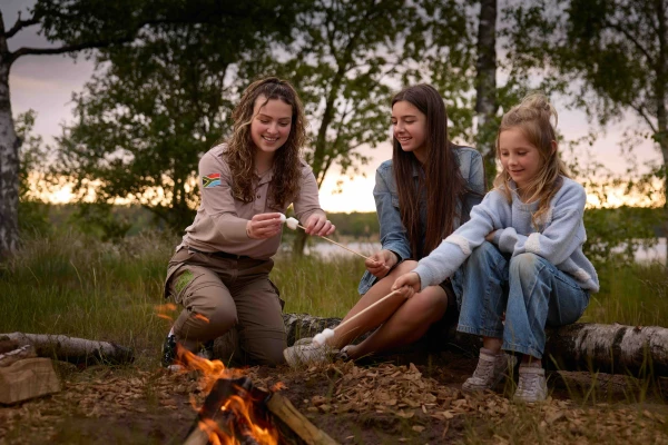 Een ranger van het animatieteam roostert marshmallows boven een kampvuur samen met twee lachende meisjes in Lake Resort Beekse Bergen