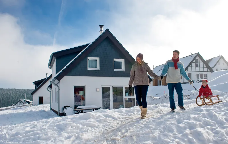 Een familie wandelt hand in hand door de sneeuw naar hun vrijstaande chalet op vakantiepark Landal Winterberg, met een kind op een slee.