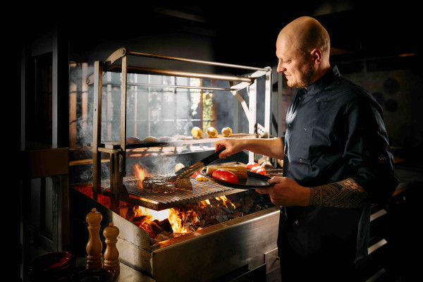 Chef‑kok bereidt gegrilde groenten en vlees op grote open houtskoolgrill tijdens live cooking diner in restaurant van Safari Resort Beekse Bergen