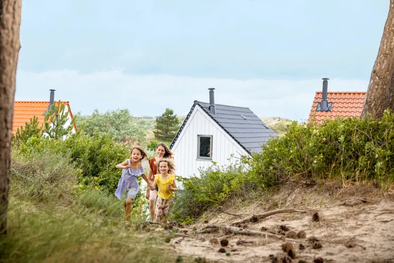 Drie kinderen rennen lachend door het duinlandschap op Center Parcs Zandvoort, met de cottages op de achtergrond.