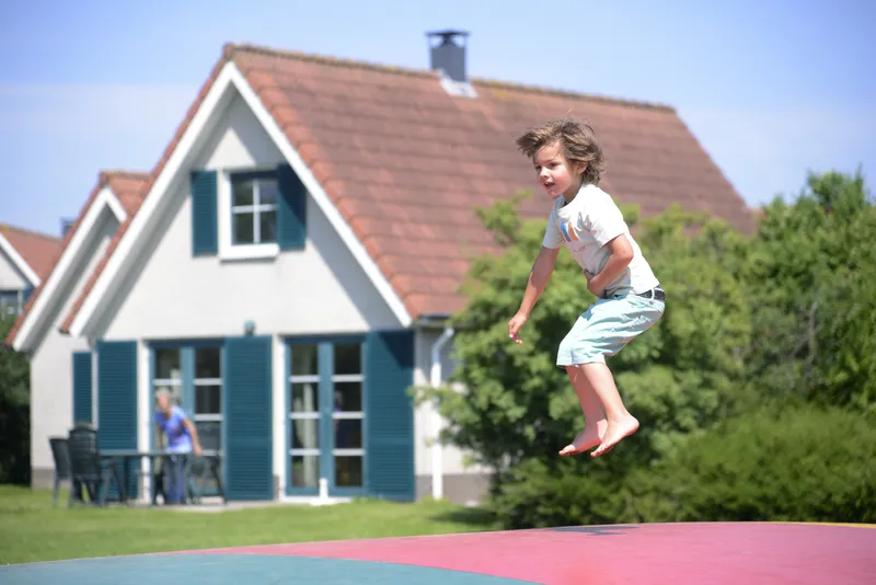 Een jongen springt hoog in de lucht op de grote air trampoline, met een comfortabel vakantiehuis van Vakantiepark Sandur op de achtergrond.